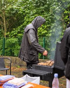 Trotz des Regens wird tapfer gegrillt. Ein Mann mit Regenjacke wendet Grillgut auf dem Rost.