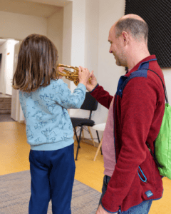 Ein Kind versucht sich am Trompete spielen. Der Vater hilft ihr dabei.