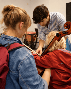 Gabriele Riccucci hilft einem Kleinkind den Cello-Bogen zu halten und eine Seite anzuspielen. Die Mutter des Kindes sitzt daneben und hält das Instrument zusätzlich fest.