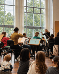 Das Göttinger Symphonieorchester im Probensaal. Nicholas Milton dirigiert. In Vordergrund sitzen Kinder auf dem Boden, sehr nah an den Orchestermusikern.